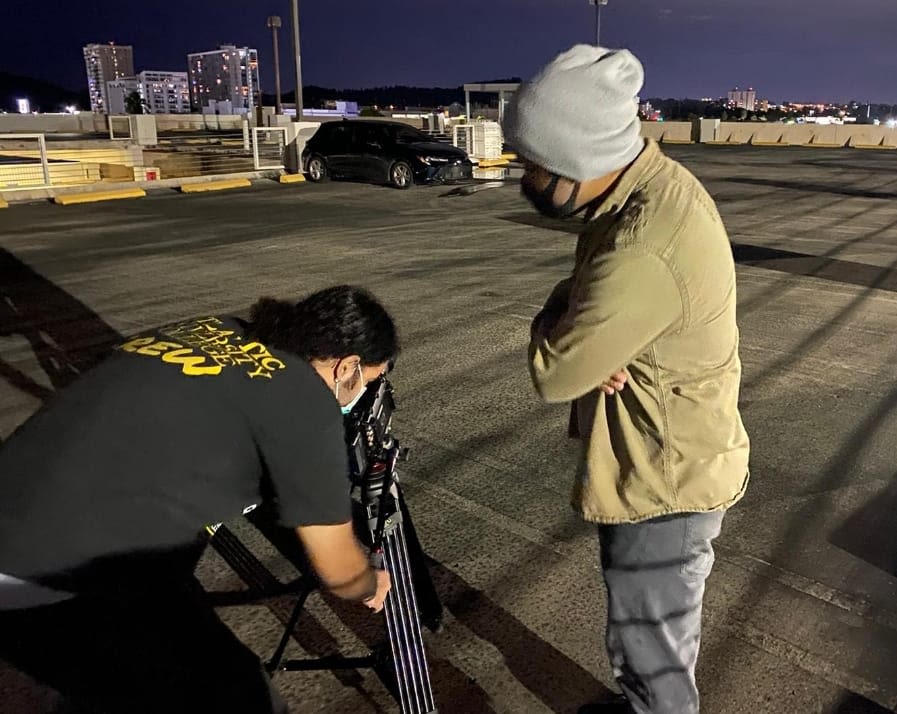 Camarógrafo con camisa negra inclinado mirando por el lente de su cámara. A su derecha observándolo está parado el director Derick Acosta con un jacket crema.