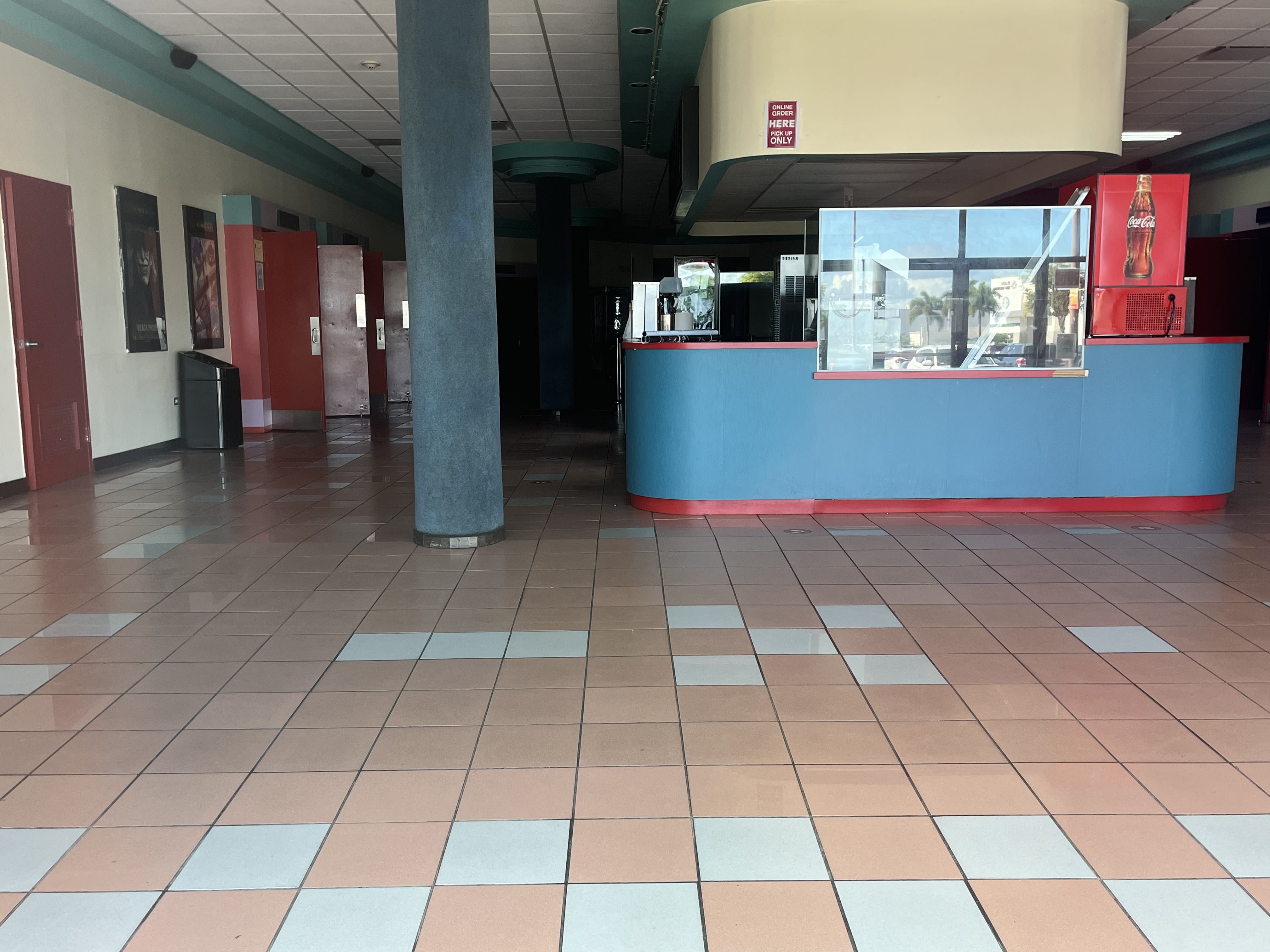 En la foto se ve una concesión de refrigerios en el lobby de un cine vacío. A mano izquierda de la foto, hay una puerta roja cerrada y tres puertas rojas abiertas. Al fondo del counter, el pasillo está oscuro.