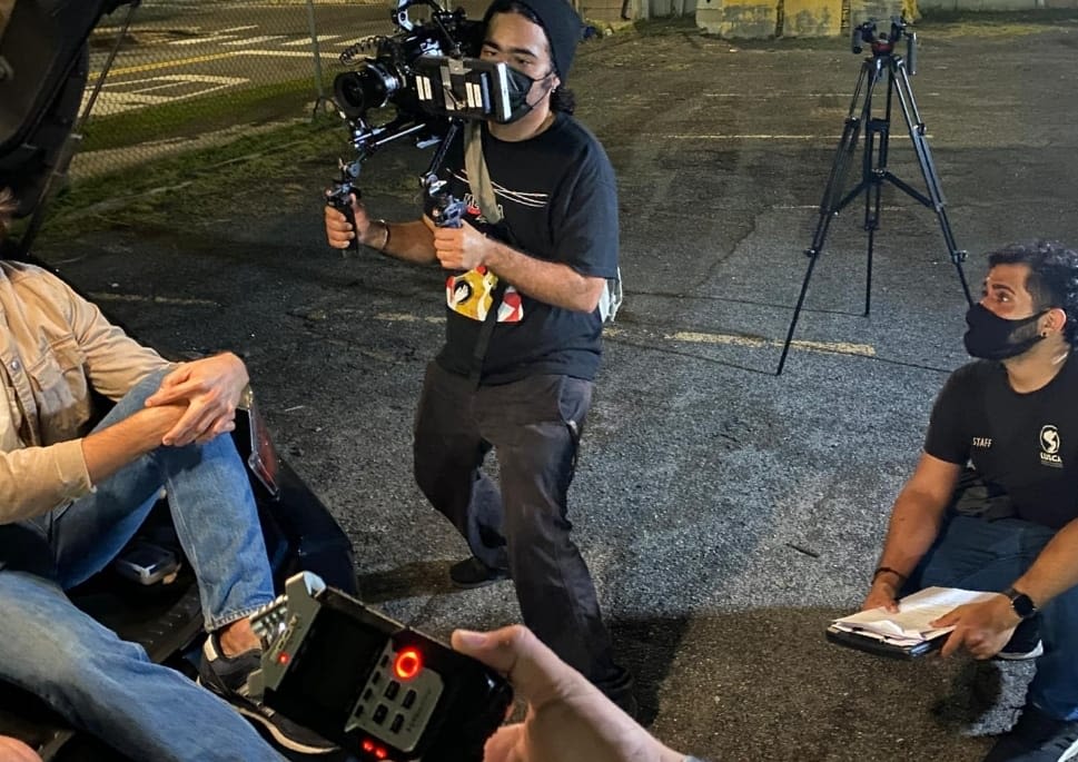 Camarógrafo con camisa negra aguantando su cámara mientras graba a uno de los actores. Sentado en el piso a mano derecha del camarógrafo está el director Derick Acosta con mascarilla y camisa negra.