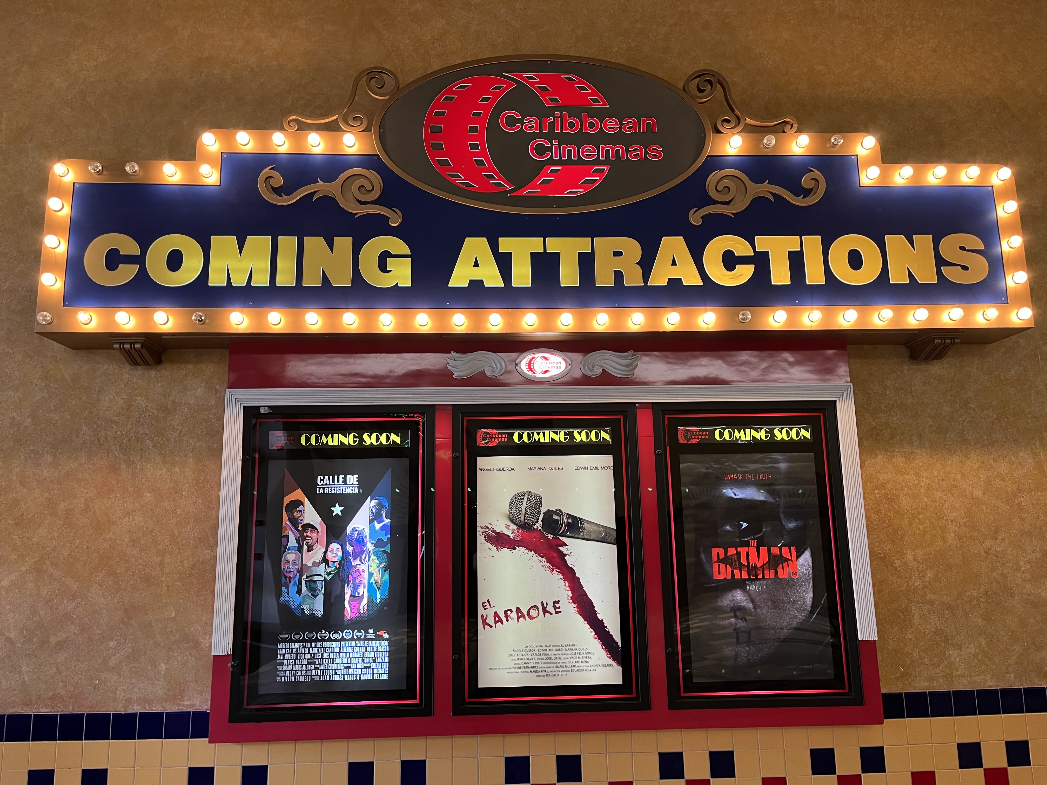 Letras amarillas rodeadas de bombillas amarillas leen "Coming Attractions". Debajo de las letras aparecen tres carteles de película. Dos de las carteleras anuncian películas puertorriqueñas: "Calle Resistencia" tiene un fondo negro y varios colores en una silueta de la bandera de Puerto Rico; y "El Karaoke" lleva un fondo blanco y un micrófono roto y sangriento. La tercera cartelera es oscura y dice "Batman"