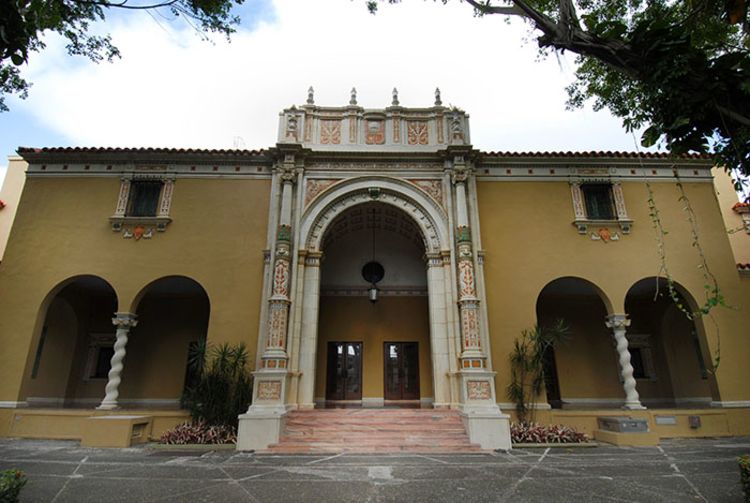 En la foto se ve la fachada principal del teatro de la UPRRP. El edificio color mostaza tiene dos arcos medianos a cada lado de su fachada de al frente. La entrada principal, que está entremedio de los cuatro arcos, tiene unas escaleras color tierra al frente. En el fondo, se ven dos puertas de madera.