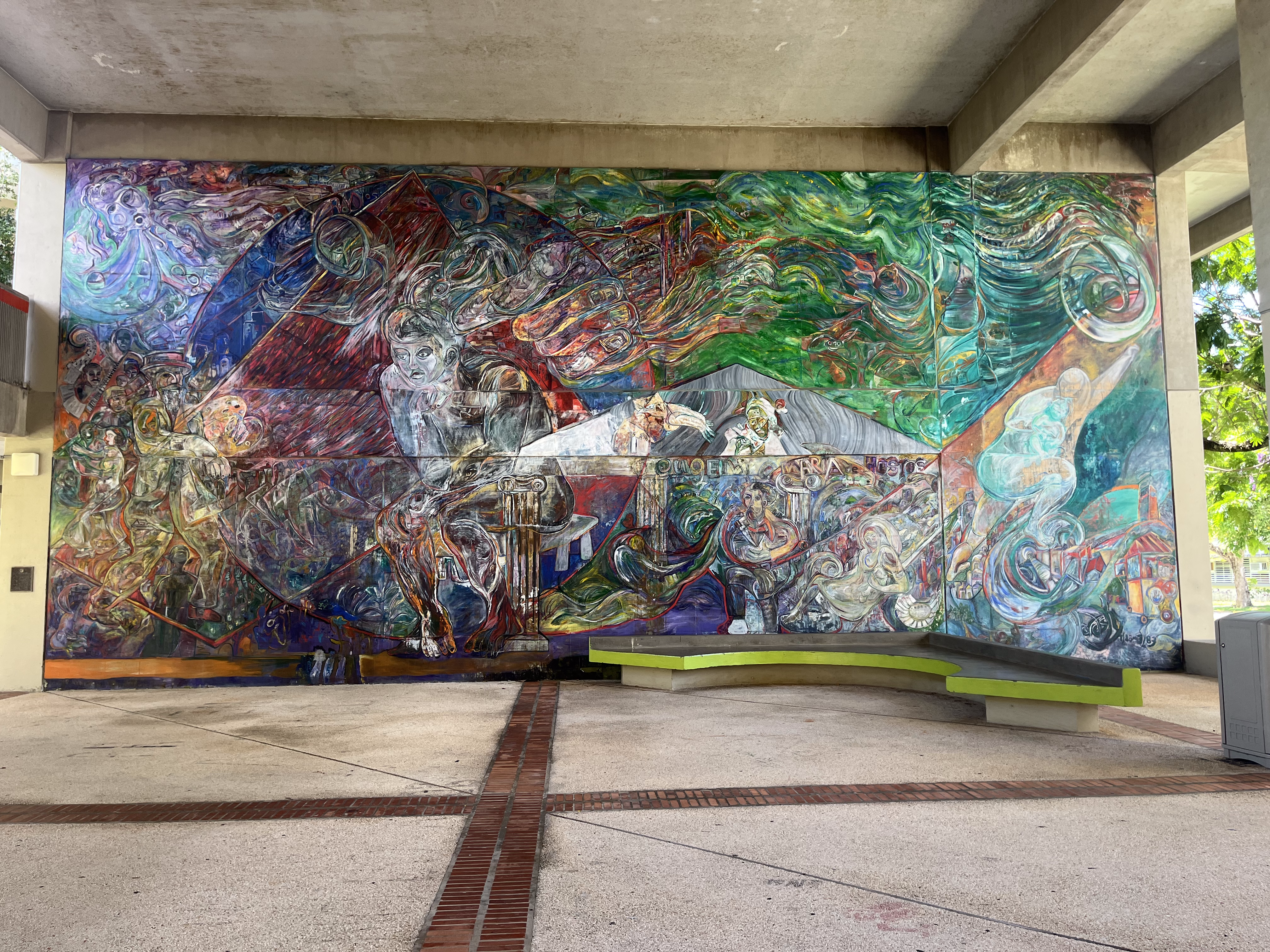 La foto muestra la obra de arte en la pared del primer piso en la facultad de Educación de la UPRRP. En el lado izquierdo del arte se ve una mezcla de los colores azul, rojo, blanco y un poco de amarillo. En el lado derecho del arte predomina el verde, blanco y un poco de crema y gris. Frente a la obra de arte, hay un banco de cemento para sentarse.